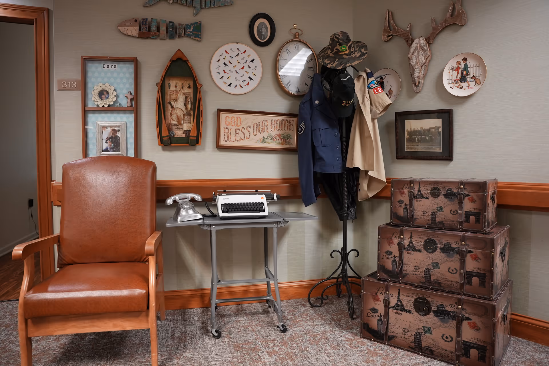 A cozy decorated interior corner with a leather armchair, a small table holding a vintage typewriter and telephone, a coat rack with jackets and hats, and stacked travel trunks beneath assorted wall decorations.