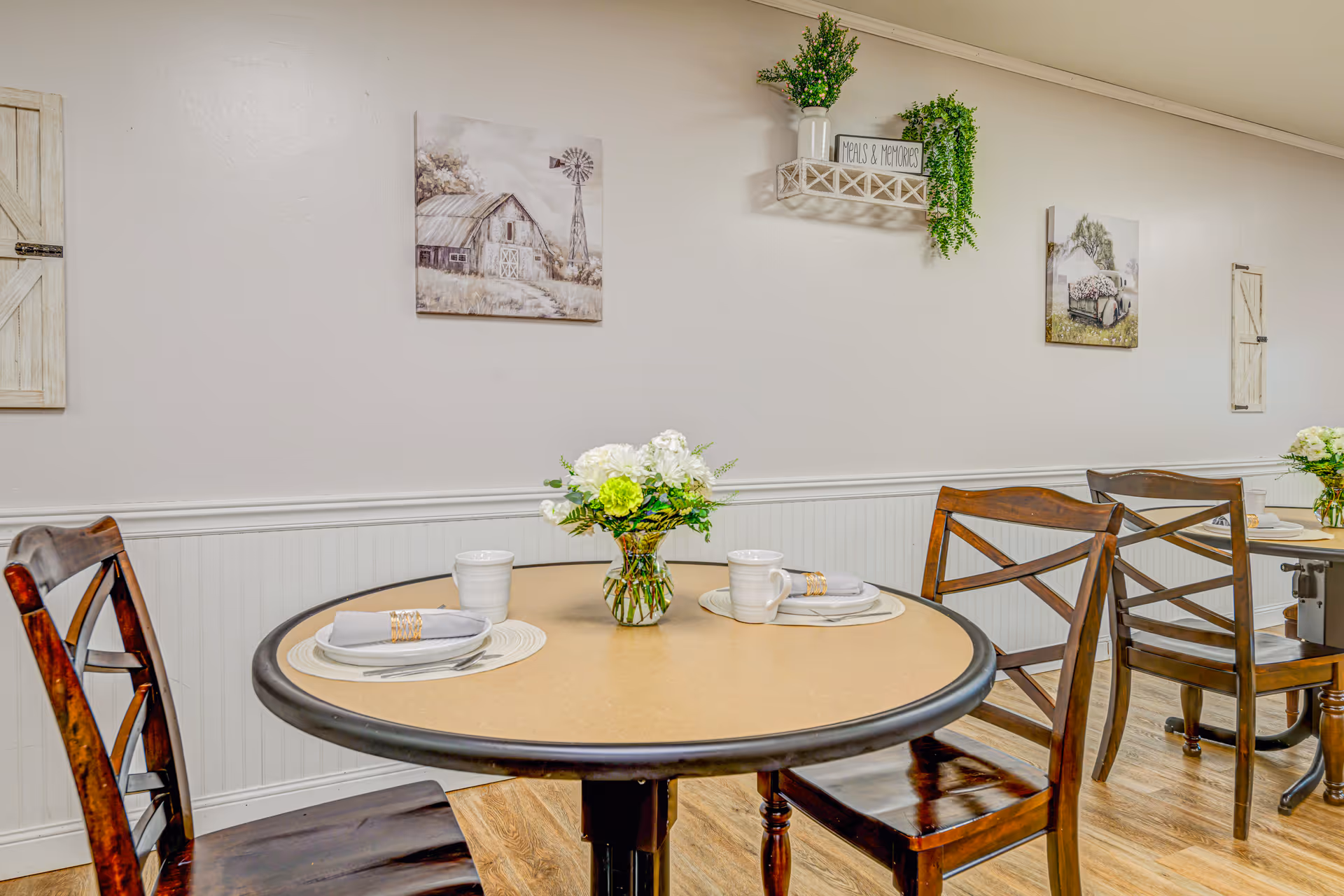 A dining area with round tables set with white plates, napkins, and cups. Each table has a small floral centerpiece with white and green flowers. The walls are decorated with rustic-themed artwork and a shelf holding a plant and a sign that reads 'Meals & Memories'. Wooden chairs surround the tables, and the floor is a light wood finish.