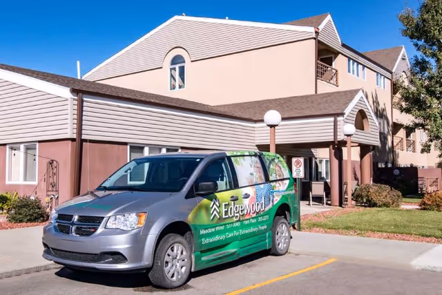 A silver and green Edgewood Park Place van parked in front of a multi-story senior living facility building with beige and brown exterior walls, a small lawn, and a clear blue sky.