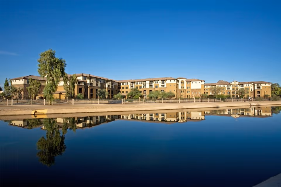 A long multi-story beige building reflected in a calm canal under a clear blue sky.