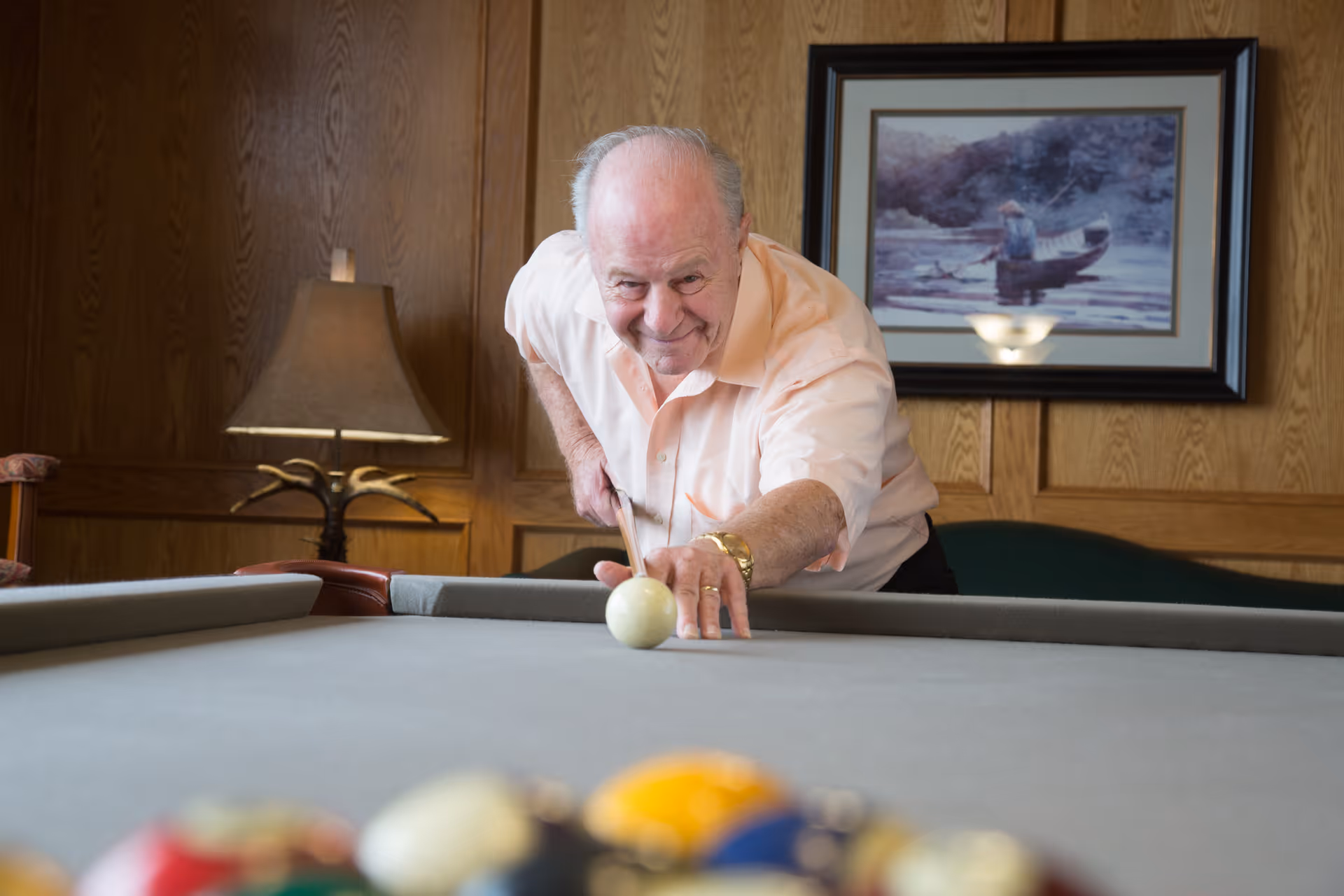 An elderly man playing pool indoors, aiming to strike the cue ball on a billiards table. The room has wood-paneled walls, a table lamp, and a framed painting of a person fishing in a boat on a lake.