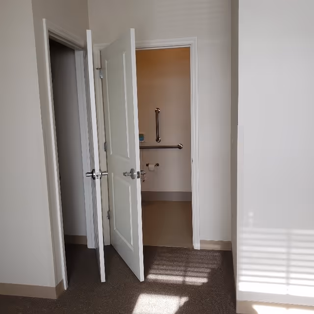 View of an interior hallway with two open white doors leading to a bathroom. The bathroom has beige walls, a grab bar, and a toilet paper holder visible. Sunlight casts shadows on the carpeted floor and white walls.