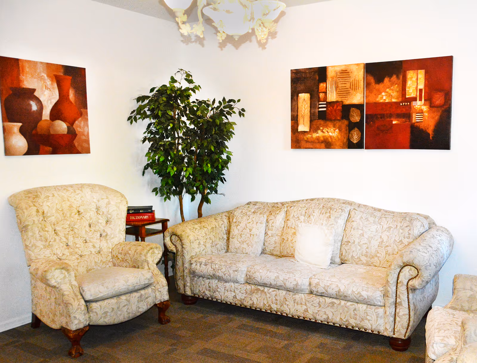 A cozy living room area with a patterned beige armchair and matching sofa. There is a small wooden side table with books on it, a tall green potted plant, and two abstract paintings with warm tones hanging on the white walls. A decorative chandelier is partially visible on the ceiling.