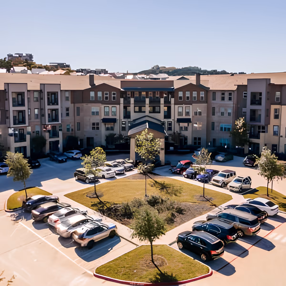 Exterior view of a multi-story senior living facility with a central courtyard featuring green grass, trees, and a circular driveway surrounded by parked cars under a clear sky.