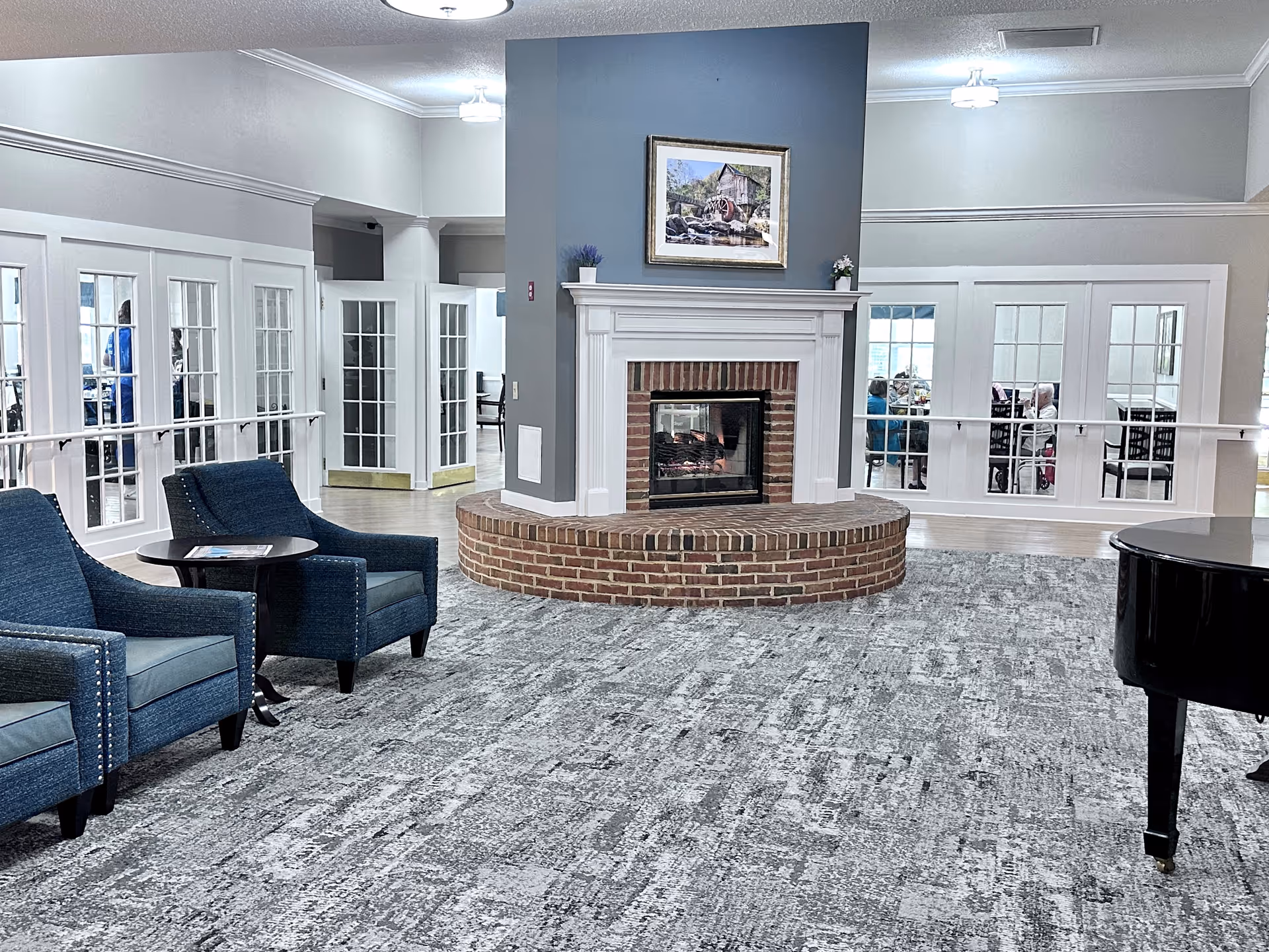 Spacious senior living common room featuring a central brick fireplace, blue upholstered chairs, and a black grand piano.
