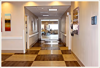 Well-lit interior corridor with patterned wood flooring leading to a common area visible through a glass opening.