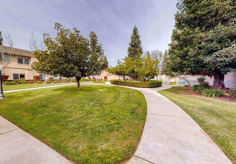 Curved concrete pathways winding through a well-maintained grassy courtyard with several trees and shrubs, surrounded by a two-story building under a cloudy sky.