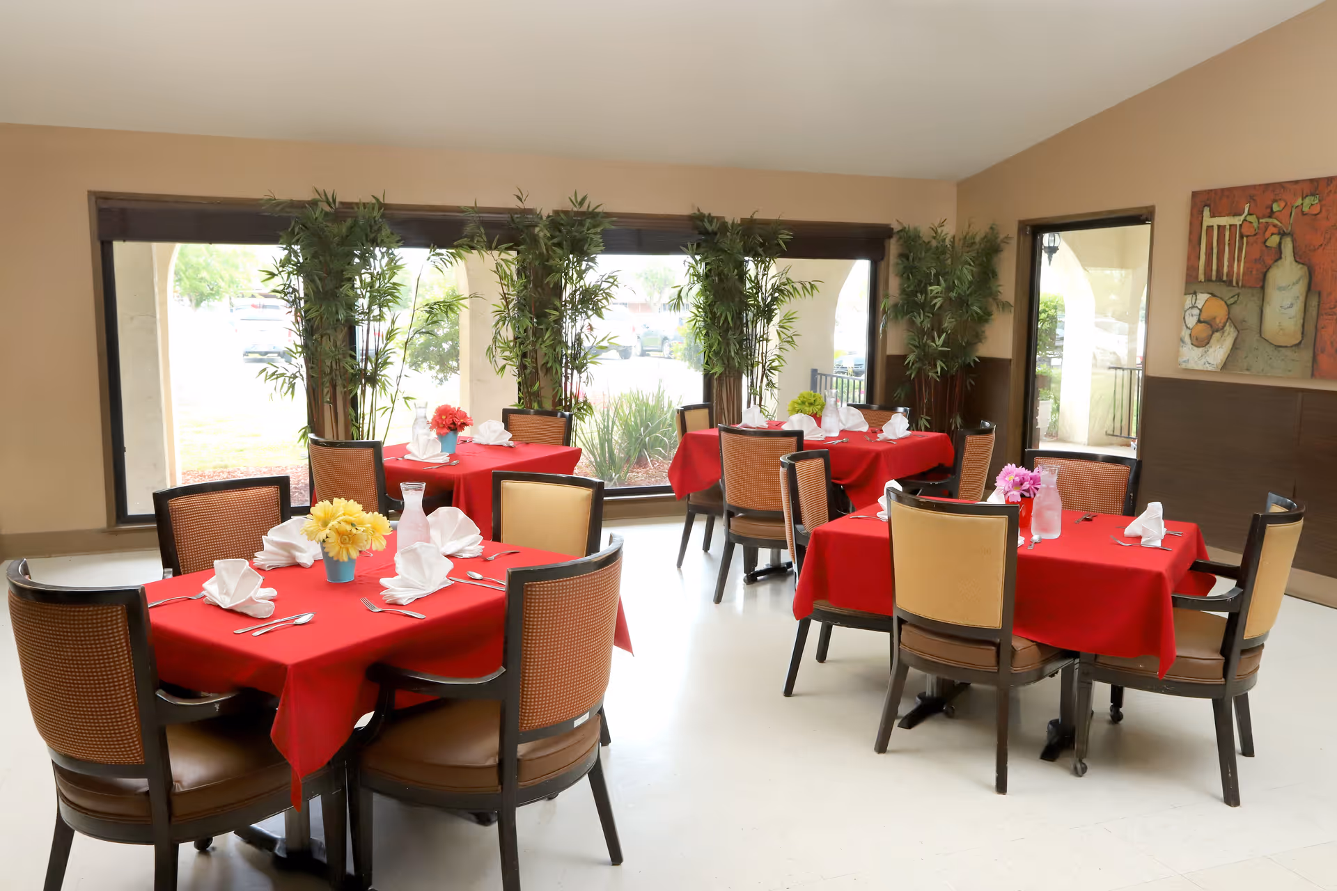 Bright dining room with several tables draped in red tablecloths, set with folded napkins and floral centerpieces, surrounded by chairs and large windows with potted plants.