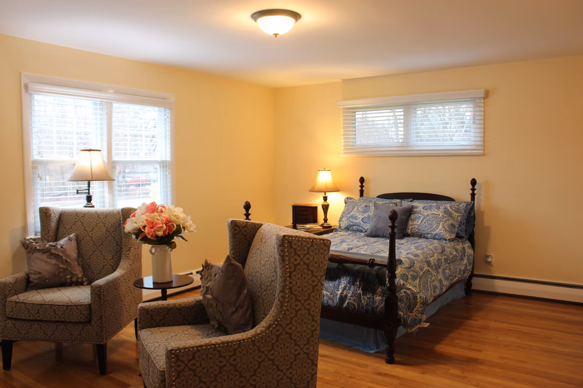 A cozy bedroom with a wooden bed featuring blue patterned bedding and a gray pillow. Two upholstered armchairs with decorative pillows are positioned near a window with white blinds. A small round table with a vase of pink and white flowers sits between the chairs. Two table lamps provide warm lighting, and the room has light yellow walls and hardwood flooring.