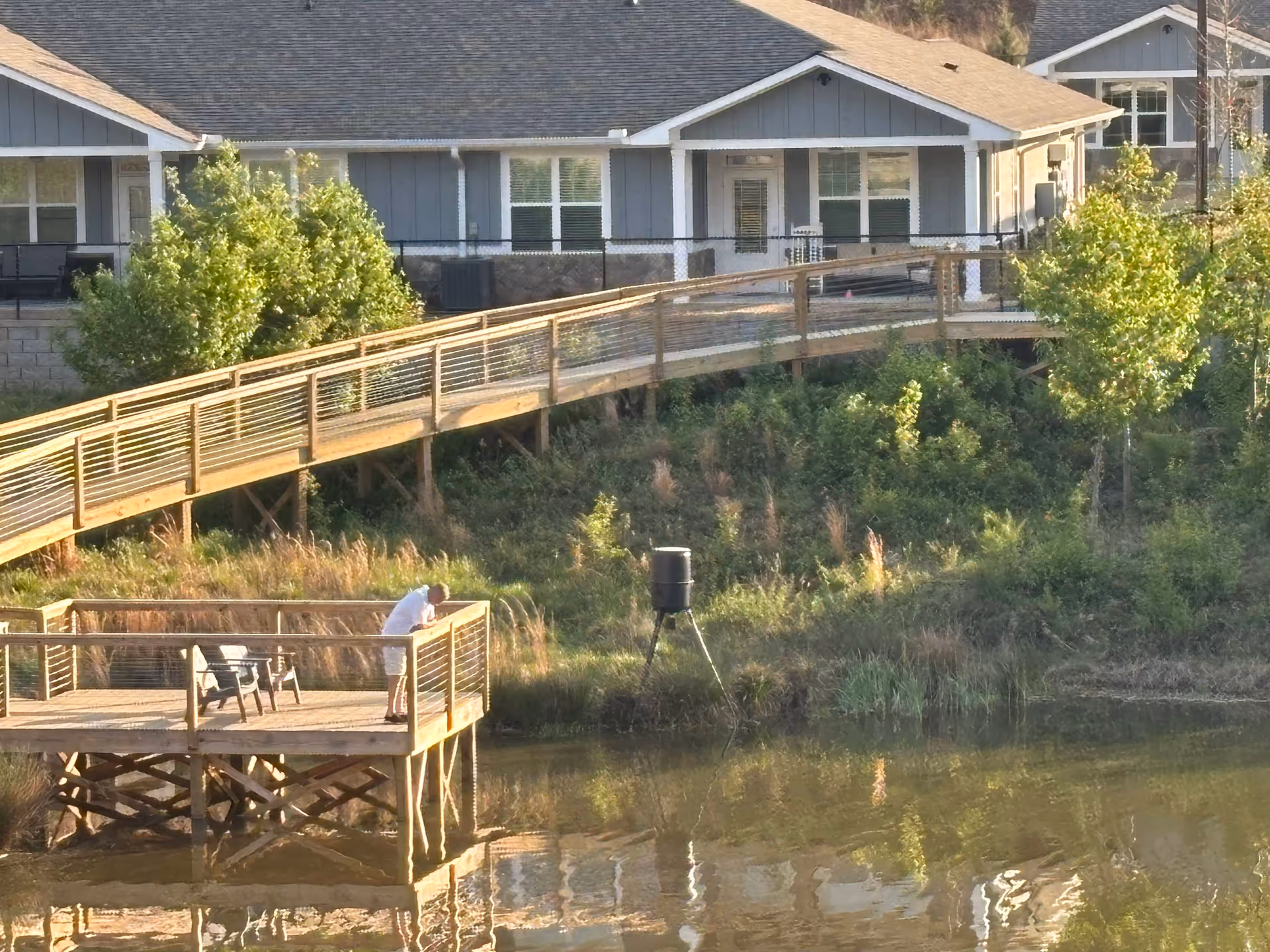 A person standing on a wooden dock overlooking a pond with a house and a wooden ramp in the background surrounded by greenery.