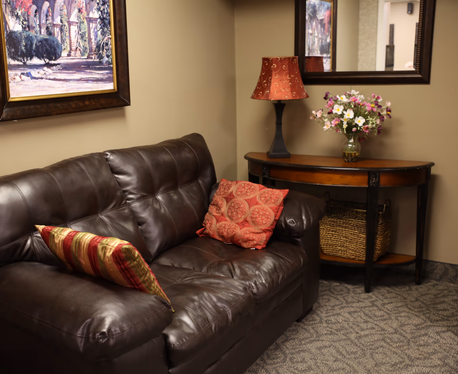 Leather couch with patterned throw pillows beside a console table holding a lamp and a vase of flowers in a cozy interior.