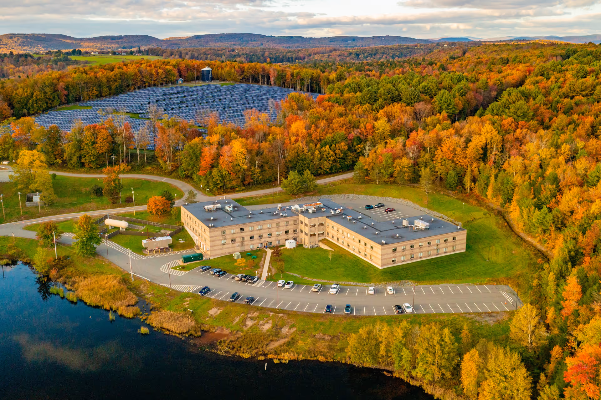 Aerial view of a large, three-story care facility building surrounded by autumn-colored trees and a parking lot with several cars. The building is situated near a body of water, with a backdrop of rolling hills and a solar panel installation on a hillside.