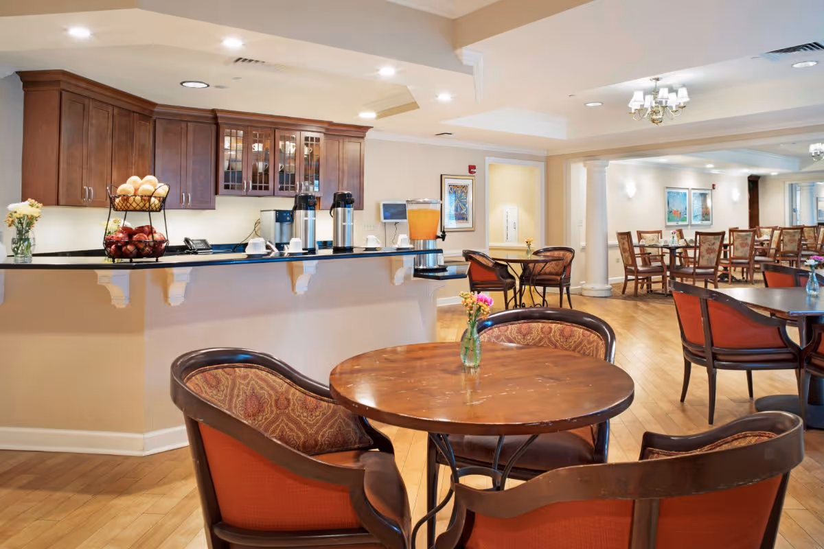 Bright communal dining area with round wooden tables and upholstered chairs, and a serving counter with coffee urns and a juice dispenser.