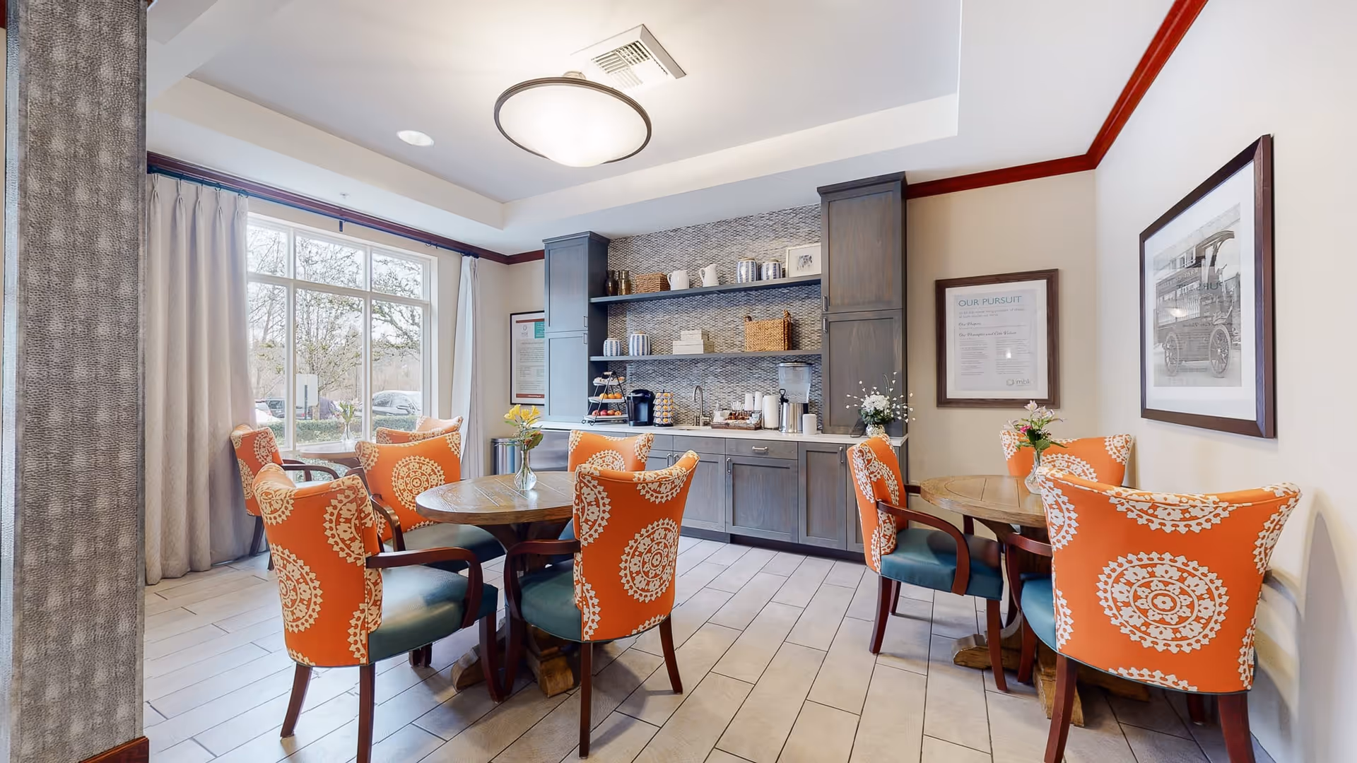 A bright dining area with two round wooden tables surrounded by orange chairs with white circular patterns. The room features large windows with curtains, a ceiling light fixture, and a built-in cabinet with shelves holding coffee supplies and decor items.