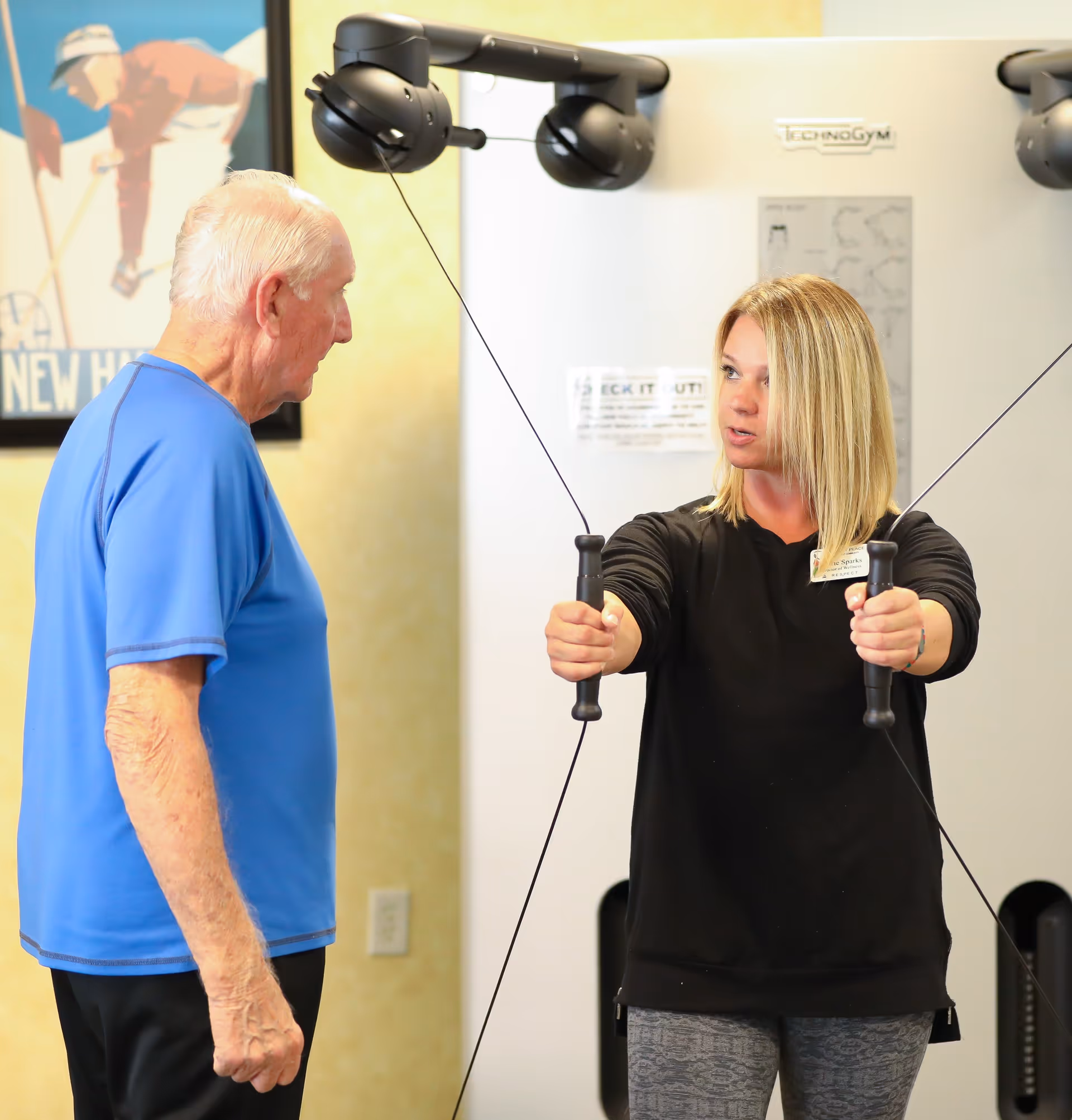 An elderly man in a blue shirt is receiving guidance from a female fitness instructor who is demonstrating an exercise using a cable machine in a gym setting. The instructor is holding the handles of the machine with arms extended, while the man watches attentively. A framed sports poster is visible on the wall in the background.