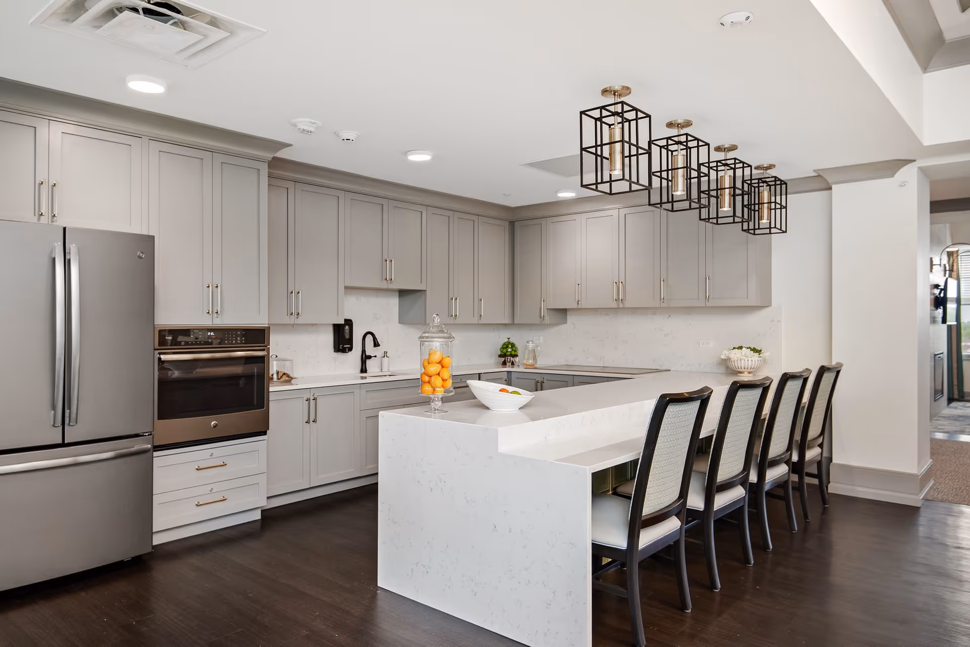 Modern kitchen with gray cabinets, stainless steel refrigerator and oven, white marble countertops, a large island with seating for five, and decorative pendant lights hanging above the island.