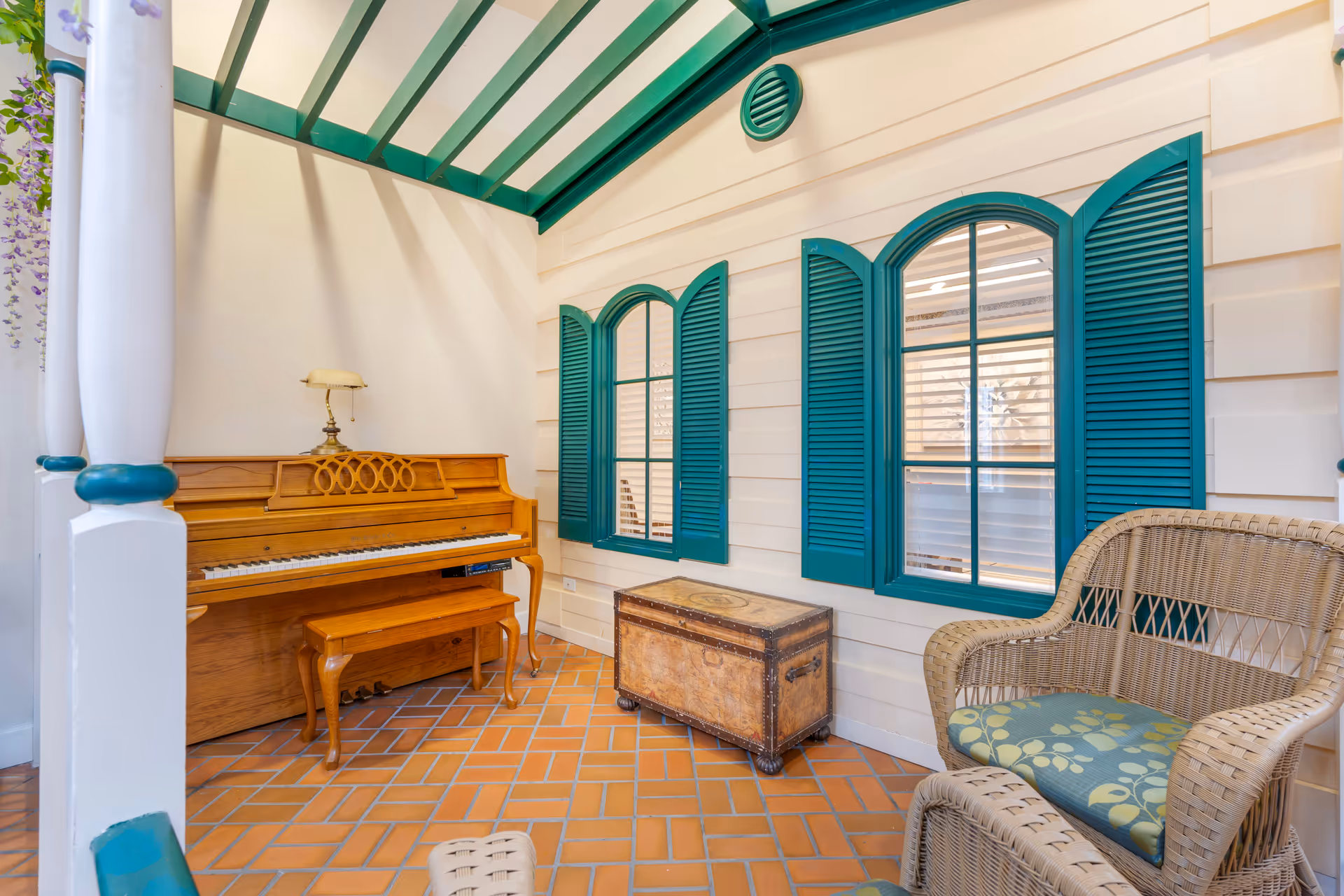 A cozy indoor seating area with a wooden piano and matching bench, a vintage trunk, and wicker chairs with green patterned cushions. The walls are white with teal window shutters and a teal-framed glass ceiling allowing natural light to enter.