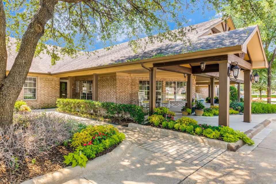 Exterior view of Anointed Senior Living featuring a landscaped entrance with colorful flowers, a covered porch with rocking chairs, and large windows.