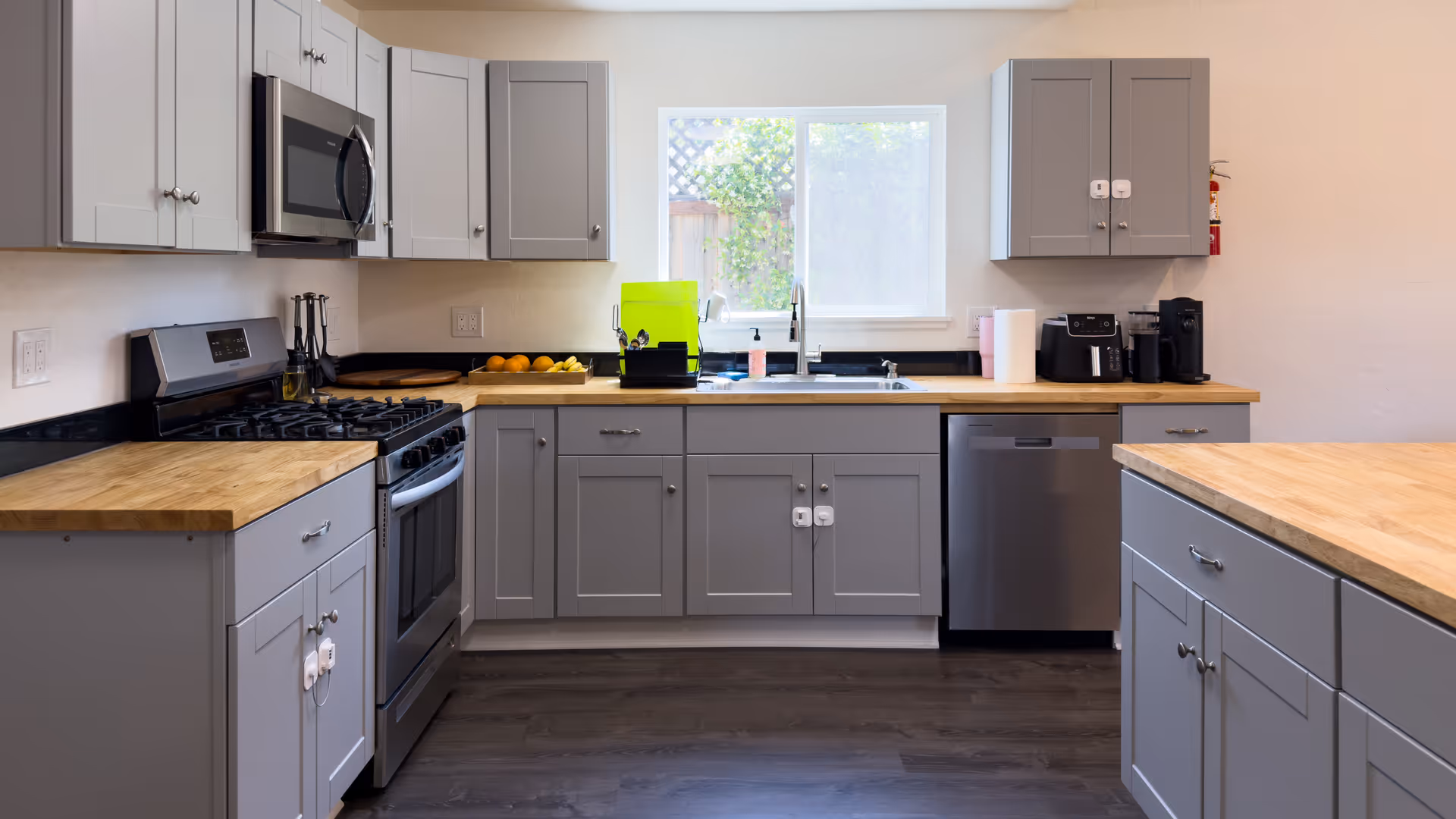 Bright modern kitchen with gray cabinets, wood countertops, stainless steel appliances, and a sink beneath a window.