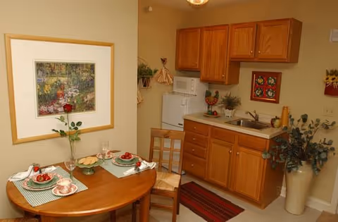 Small kitchen area with wooden cabinets, a white microwave on top of a white refrigerator, a sink, and a countertop. A round wooden dining table set for two with plates of food, cups, and a vase with a single red rose. Wall decorations include a framed picture and a small hanging ornament. A potted plant is placed on the floor near the cabinets.