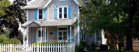 A two-story house with a white picket fence and front porch, surrounded by trees.