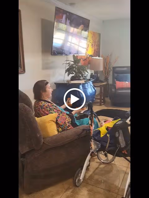 An elderly woman sitting comfortably in a brown recliner chair with a yellow pillow, smiling and looking towards a television mounted on the wall in a cozy living room. There is a blue chest with a plant and a lamp on it, a side table with decorative items, and a walker with a bag attached in front of her. The room has tiled flooring and light-colored walls.