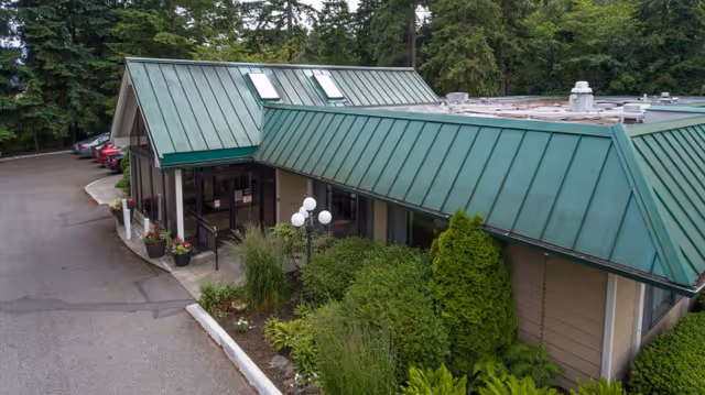 Exterior view of a single-story building with a green metal roof surrounded by trees and shrubs. There is a driveway with parked cars on the left side and a main entrance with glass doors and potted plants near the front.