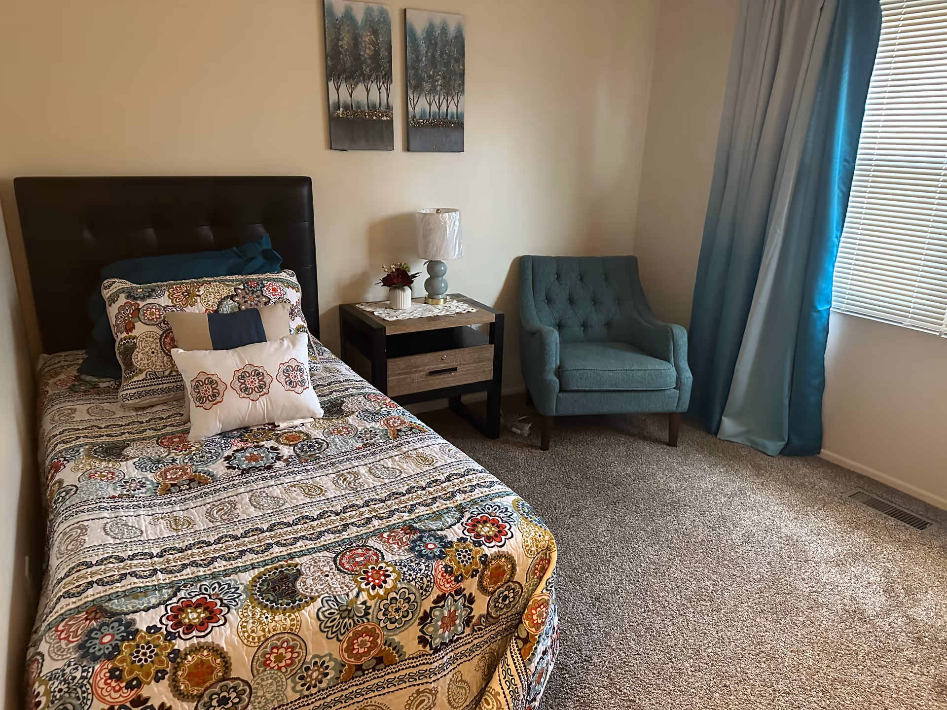 A bedroom with a single bed covered in a colorful patterned quilt and multiple pillows. Next to the bed is a wooden nightstand with a lamp and a small flower arrangement. A teal upholstered armchair is placed near a window with teal curtains and white blinds. The room has beige walls and carpeted flooring.