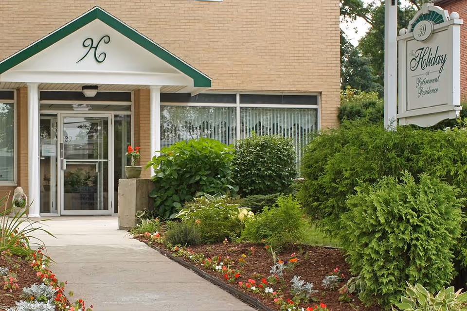 Entrance to a senior living facility with a glass door under a white and green awning featuring the letter H. There are well-maintained flower beds and shrubs lining the walkway leading to the door. A sign on the right reads 'Holiday Retirement Residence' with the number 39 above it.
