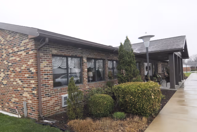 Exterior view of a single-story brick building with multiple windows covered by white curtains. There are trimmed bushes and small trees along the building, a covered entrance with a bench, and a sidewalk leading up to the entrance. The sky is overcast and the ground appears wet.