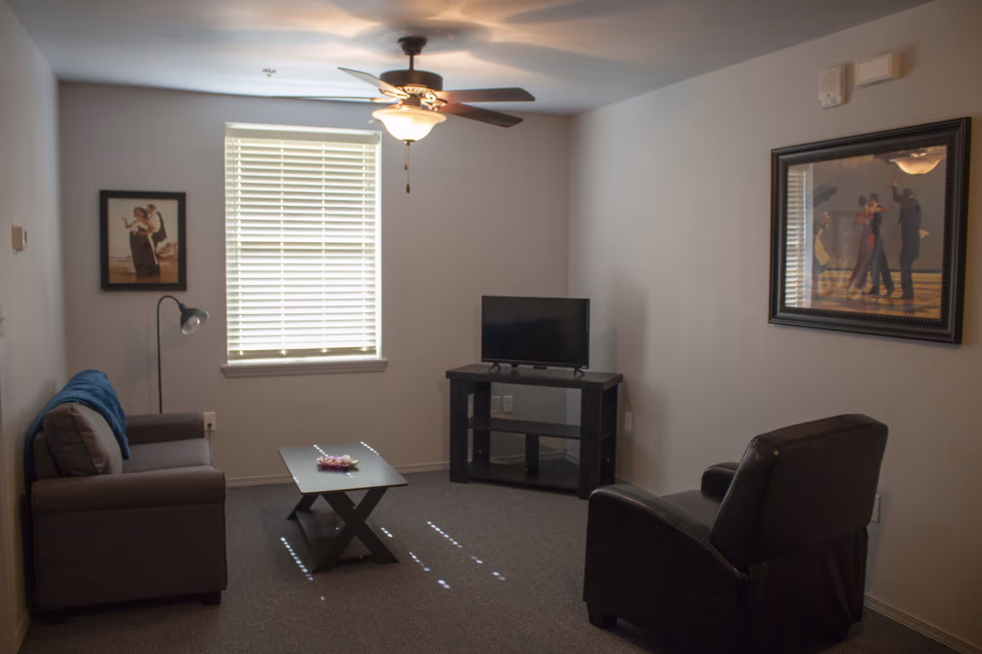 A small living room with a gray sofa on the left, a black armchair on the right, a black coffee table in the center, and a TV on a black stand against the far wall. There is a window with closed blinds in the middle of the back wall, a ceiling fan with a light fixture above, and two framed pictures on the walls.