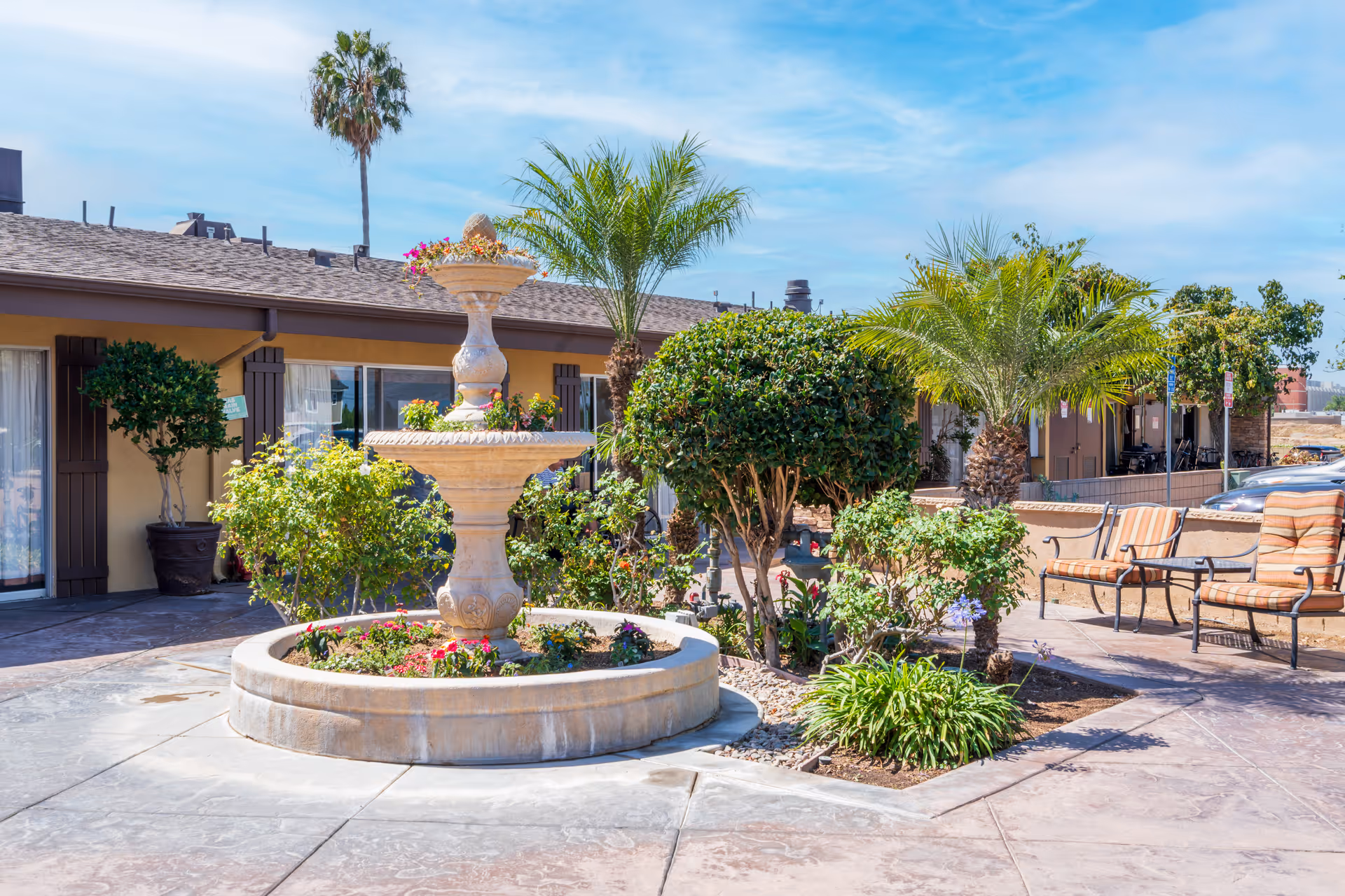 Outdoor courtyard area at Huntington Valley Healthcare featuring a stone fountain surrounded by various plants and flowers, palm trees, and patio seating with striped cushions under a clear blue sky.
