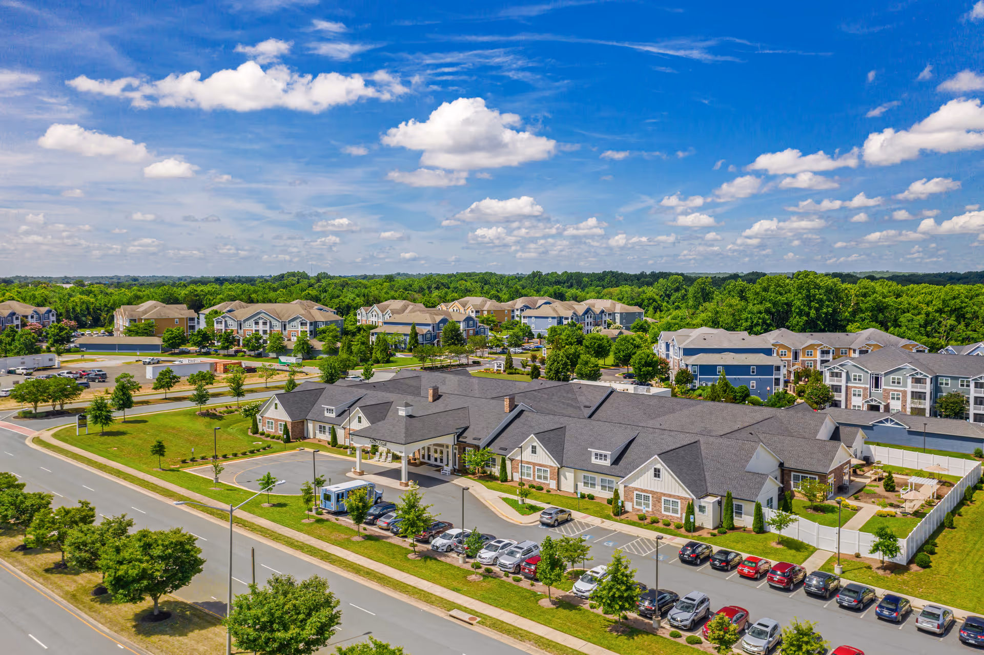 Aerial view of a single-story senior living building with parking lot, landscaped grounds, and nearby apartment complexes under a blue sky.
