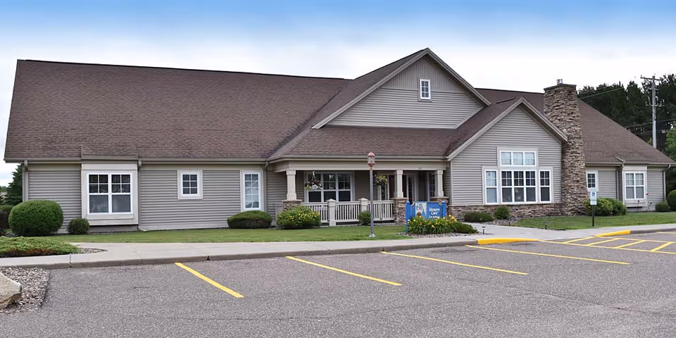 Exterior view of a single-story senior living facility building with beige siding, a brown roof, and a stone chimney. The building has several windows and a covered entrance with a small porch. There is a parking lot with marked spaces in front of the building and some landscaping with bushes and flowers near the entrance.
