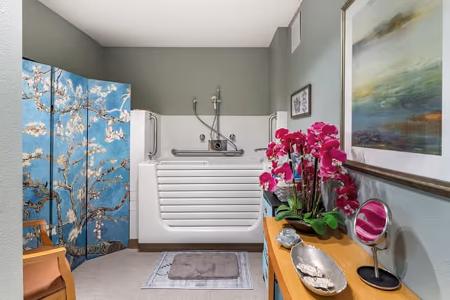A bathroom with a walk-in bathtub featuring safety rails and a handheld showerhead. The room has a decorative blue folding screen with a floral design, a wooden chair, a wooden table with pink orchids, a small mirror, and decorative items. The walls are painted light green and there is a framed painting hanging above the table.
