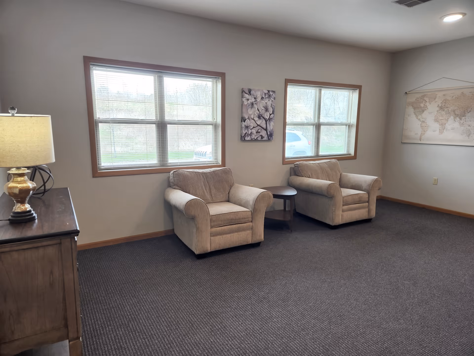 A cozy sitting area with two beige armchairs separated by a small round wooden table, positioned in front of two large windows with wooden frames. A wooden sideboard with a lamp is on the left side, and a world map hangs on the right wall. The room has gray carpet and neutral-colored walls.