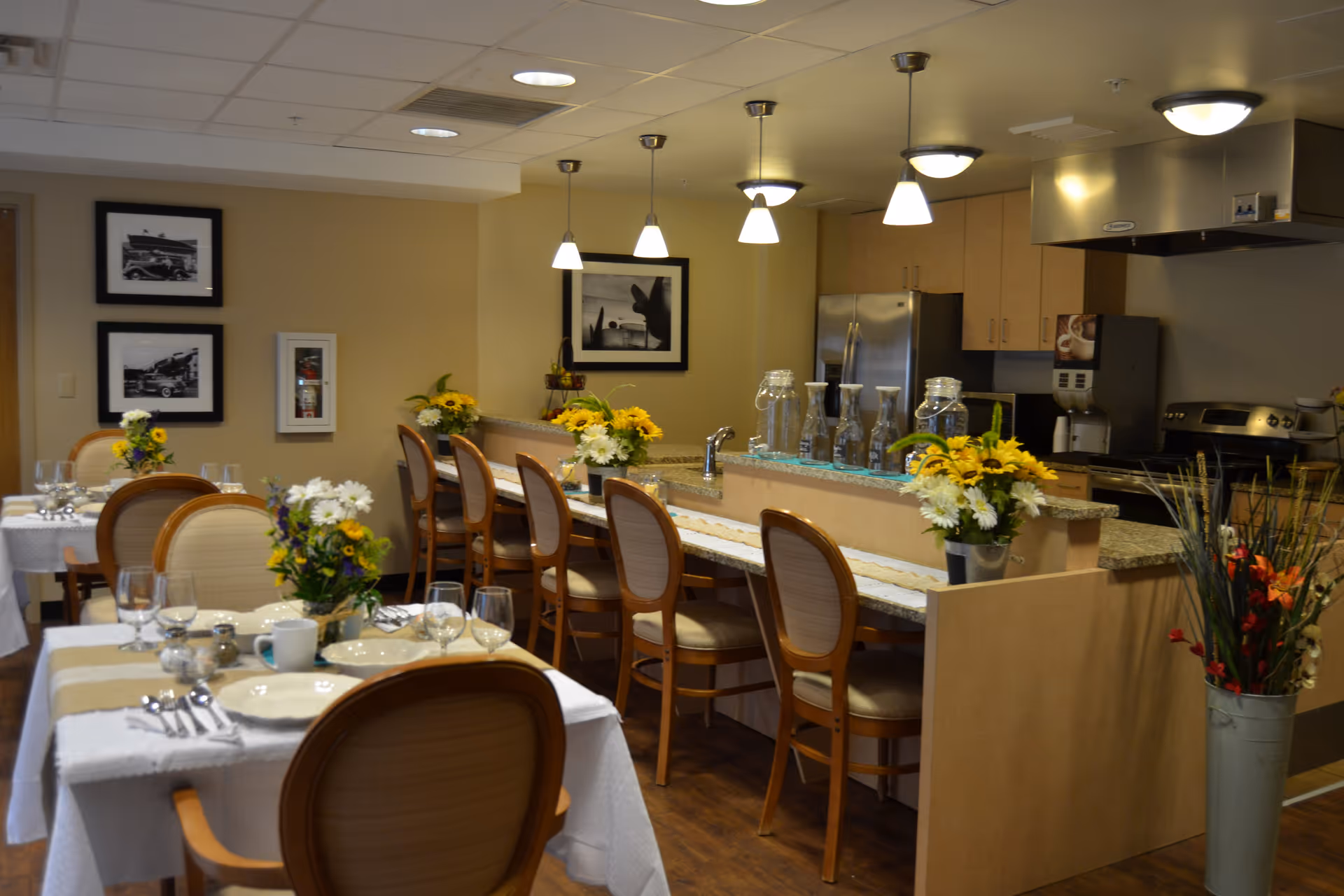 A dining area with tables set for a meal, including plates, glasses, and cutlery. There are wooden chairs around the tables and a counter with bar stools. The counter has decorative flowers and glass jars. The background shows a kitchen area with stainless steel appliances and cabinets. The room is warmly lit with ceiling lights and pendant lamps.
