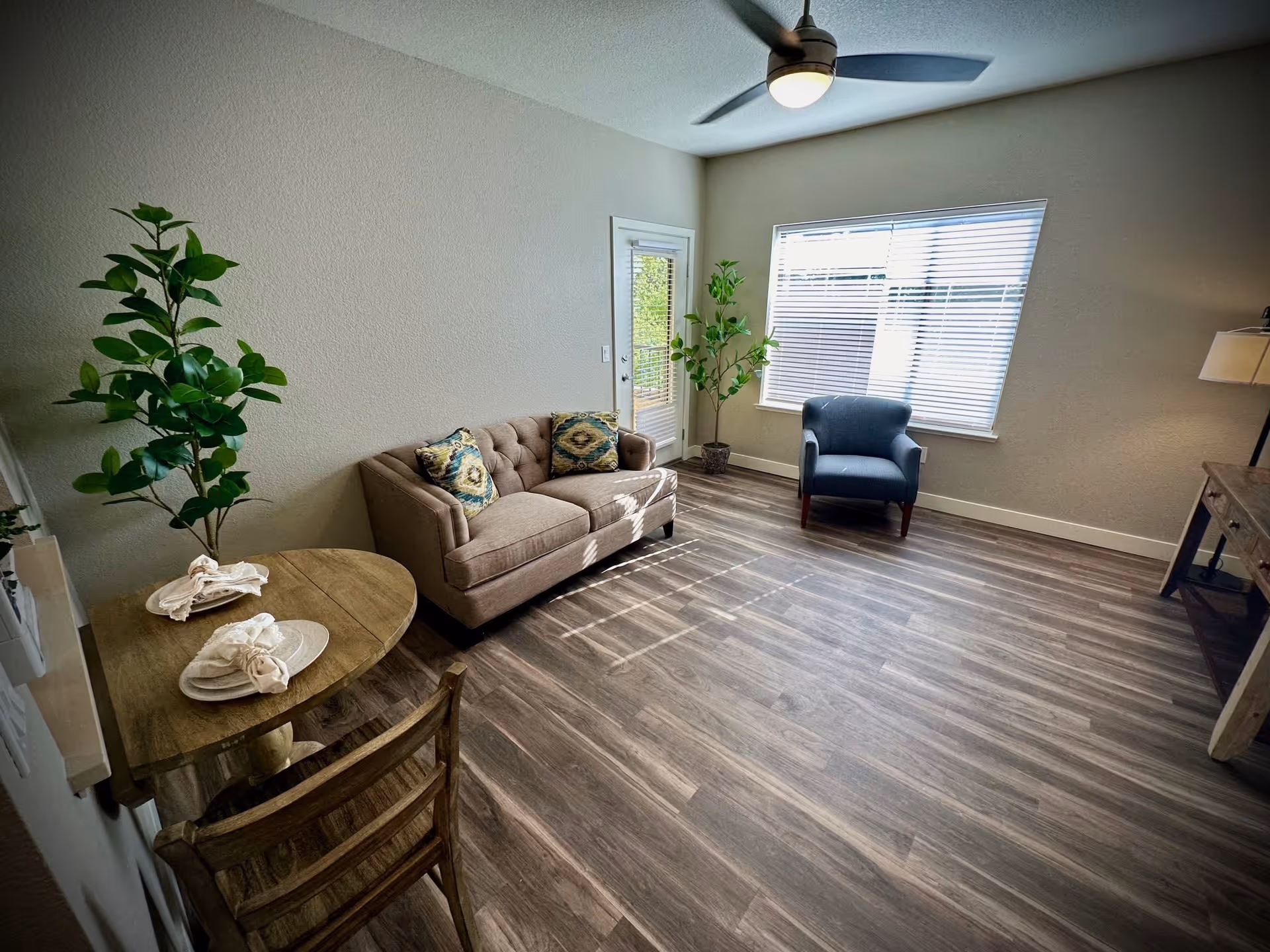 A cozy living room with a beige tufted sofa adorned with two patterned cushions, a single blue armchair near a window with blinds, a small wooden dining table set for two with napkins and plates, two potted plants, a ceiling fan with light, and a wooden console table with a lamp on the right side. The room has wood-style flooring and light-colored walls.
