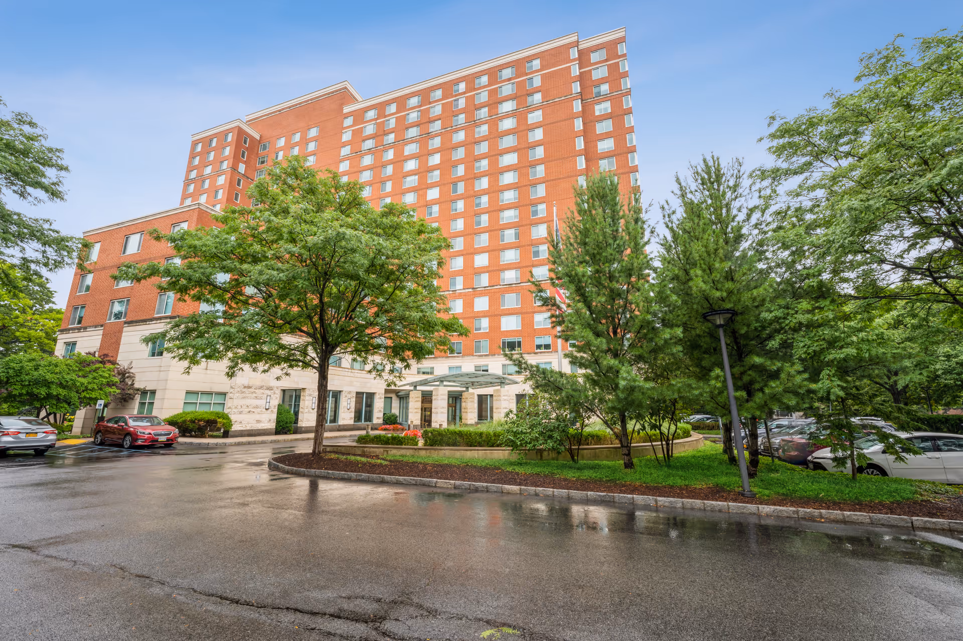 Exterior view of a multi-story brick and stone building with many windows, surrounded by trees and parked cars on a wet driveway under a cloudy sky.
