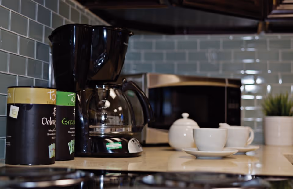 Close-up view of a kitchen countertop featuring a black coffee maker, two tins of tea, a white teapot, two white teacups on saucers, a stainless steel microwave, and a small potted plant. The backsplash is made of light green subway tiles.