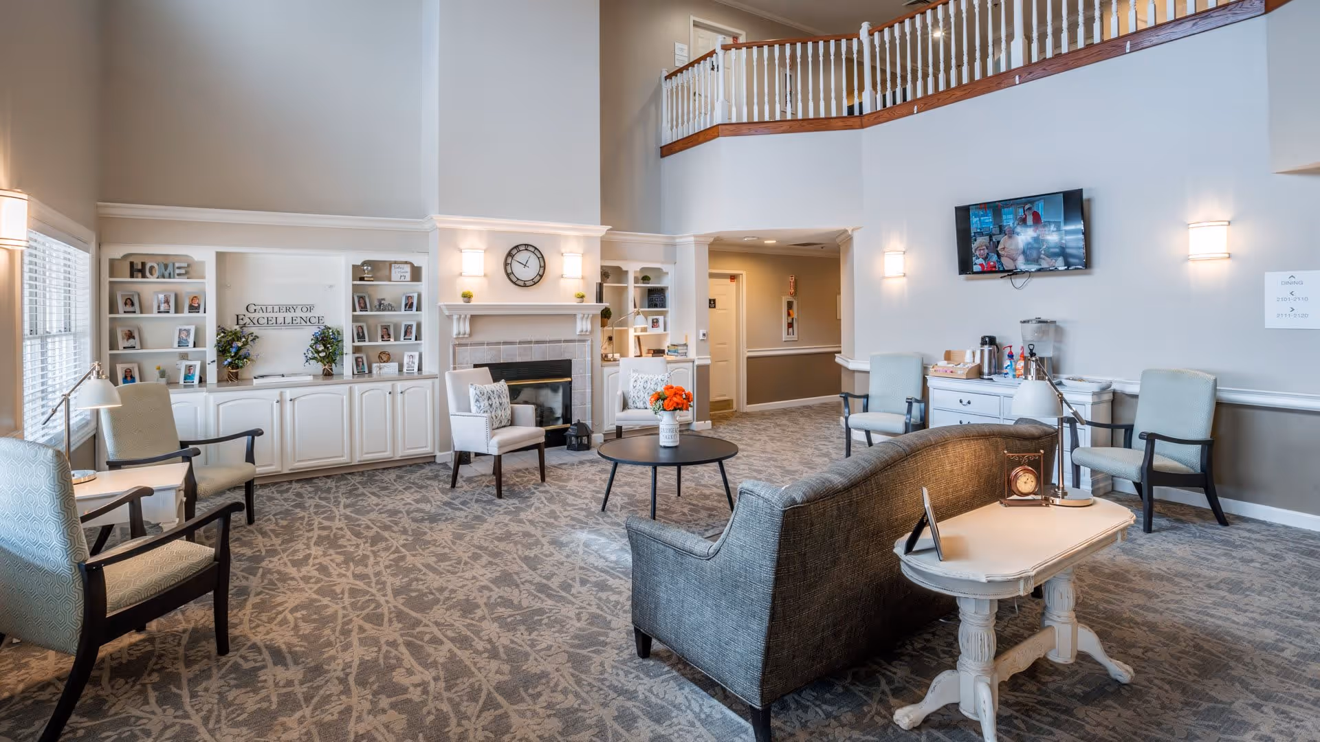 A spacious senior living facility common area with comfortable seating including armchairs and a sofa arranged around a round coffee table with a vase of flowers. There is a fireplace with a clock above it, built-in shelves with framed photos and decorations, and a TV mounted on the wall. The room has a high ceiling with a balcony railing visible above, and a carpeted floor with a patterned design.