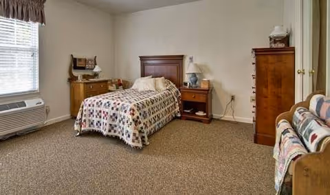 A bedroom in a senior living facility with a single bed covered in a patterned quilt, a wooden headboard, a nightstand with a lamp, a dresser, a chest of drawers, and a window with blinds and a valance. There is also an air conditioning unit below the window and a cushioned bench with a quilt draped over it.