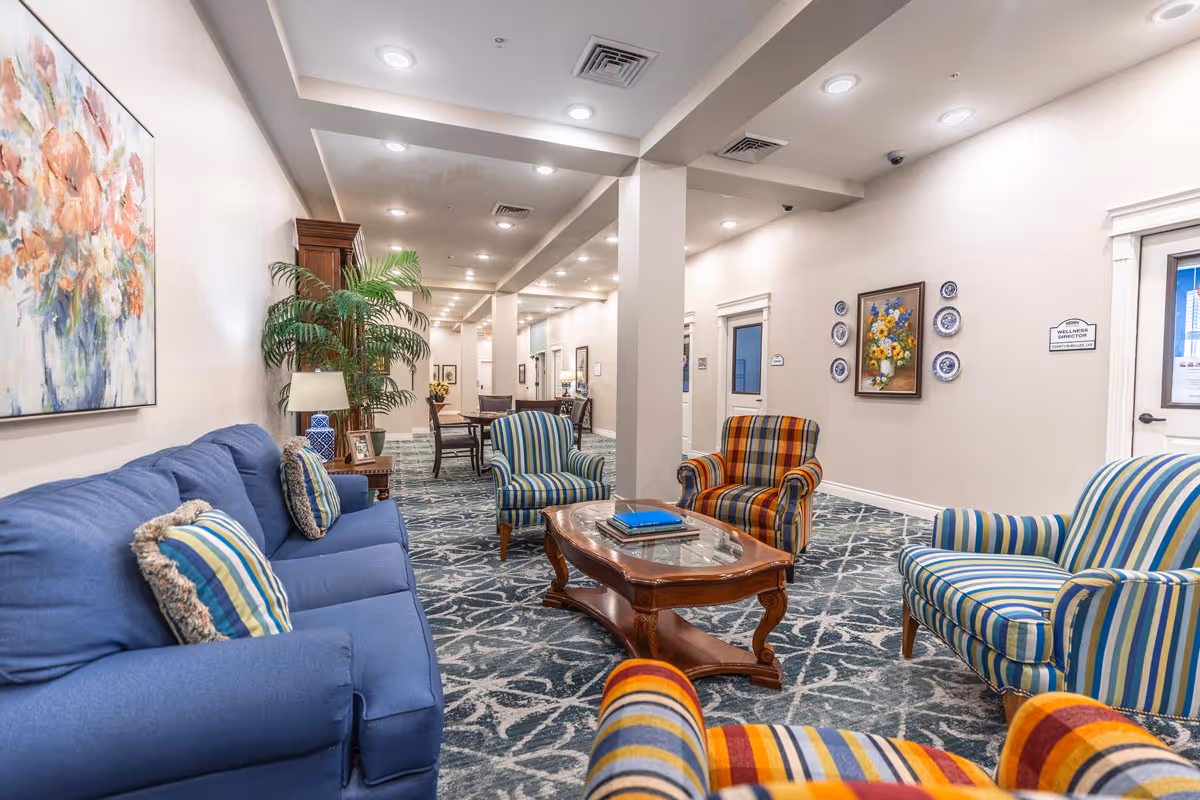 A well-lit assisted living facility common area with a blue sofa and striped armchairs in blue, yellow, and orange tones arranged around a wooden coffee table. The room features patterned carpet, framed floral artwork on the walls, potted plants, and a long hallway with additional seating and tables.