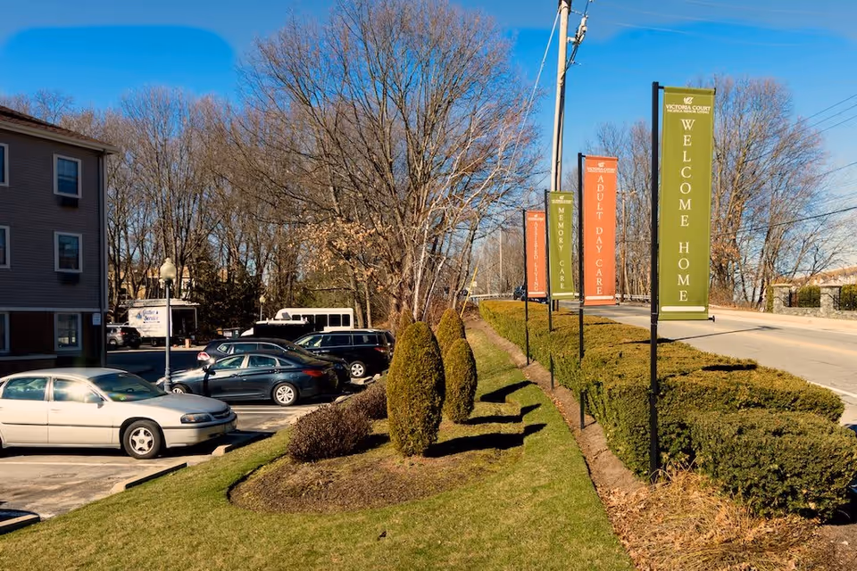 Outdoor view of a parking lot next to a road with several colorful vertical banners on poles. The banners read 'WELCOME HOME', 'ADULT DAY CARE', and 'MEMORY CARE'. There are trimmed bushes and small trees along the grassy area beside the road, and a building is visible on the left side. The sky is clear and blue.
