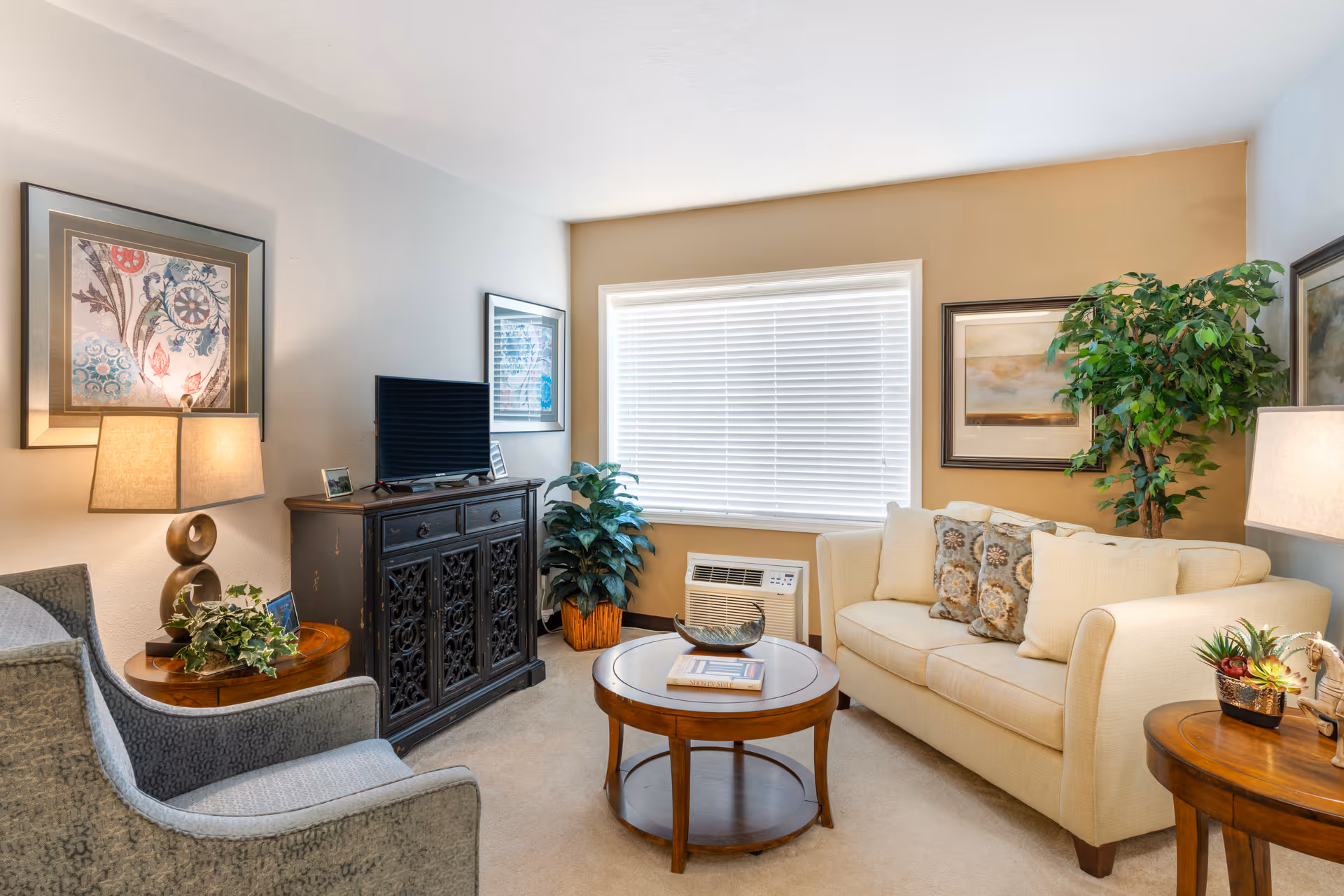 A cozy living room with a beige sofa adorned with patterned cushions, a round wooden coffee table with a book and decorative bowl, a gray armchair, two wooden side tables with lamps and plants, a black ornate cabinet with a flat-screen TV on top, framed artwork on the walls, and a large window with white blinds letting in natural light.