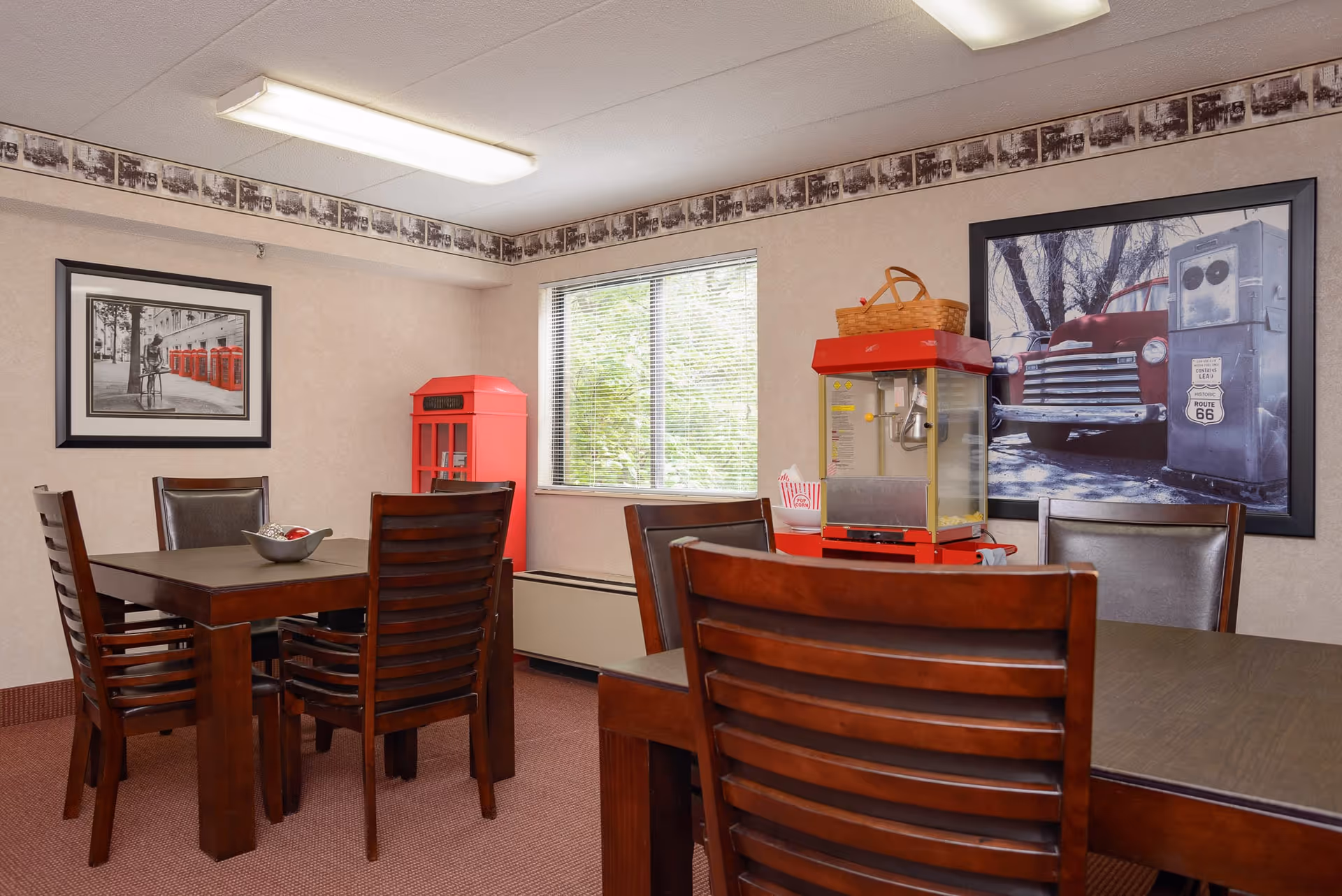 A dining area with two dark wooden tables and several matching chairs. On one table, there is a bowl with decorative items. Near the window, there is a red popcorn machine with a basket on top and a small red vintage-style phone booth decoration. The walls are beige with a wallpaper border near the ceiling featuring black and white photos. Two framed pictures hang on the walls, one showing red phone booths and the other showing a vintage red truck and a Route 66 gas pump.
