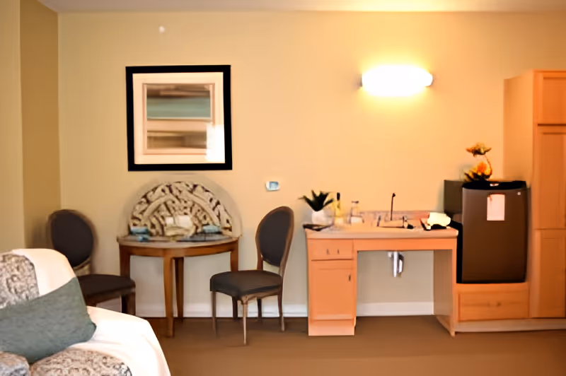 Interior view of a room with a small wooden table against a beige wall, two gray upholstered chairs on either side, a framed abstract artwork above the table, a wooden desk with a sink and faucet, a small refrigerator, and a cabinet. A couch with a white and gray patterned throw and green pillow is partially visible in the foreground.