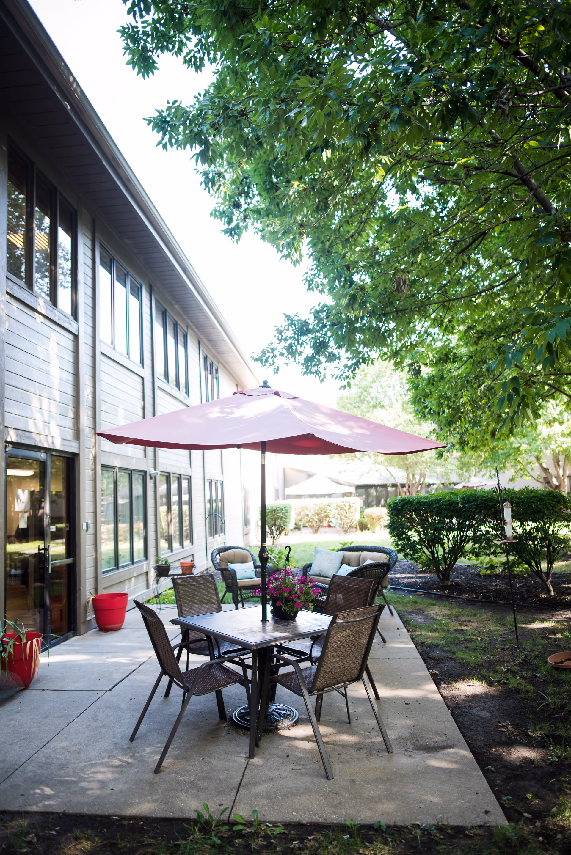 Outdoor patio area at Shawnee Gardens Healthcare and Rehabilitation Center with a table and four chairs under a large red umbrella, surrounded by greenery and bushes, adjacent to the building.