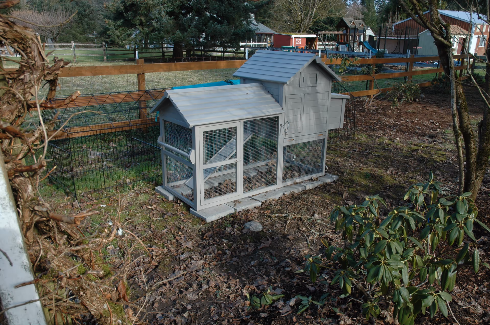 A small gray wooden chicken coop with wire mesh sides sits on a patch of ground covered with dry leaves and some green plants. The coop is placed on concrete blocks and is enclosed by a wooden fence. In the background, there are trees, a playground with slides, and some buildings.
