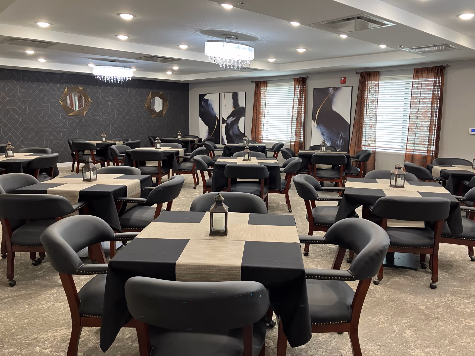 A dining room with multiple square tables covered with black and beige tablecloths, each table has a small lantern centerpiece. The room has gray cushioned chairs with wooden legs, large windows with sheer brown curtains, modern chandeliers on the ceiling, and abstract artwork on the walls.